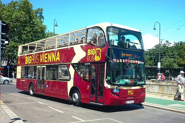 Um ônibus turístico aberto de dois andares, vermelho e creme, em uma rua arborizada de Viena, sob um céu azul