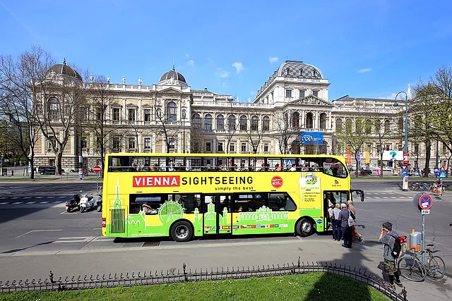 Um ônibus turístico de dois andares, amarelo e verde, na Ring diante do edifício principal da Universidade de Viena, em um dia de sol