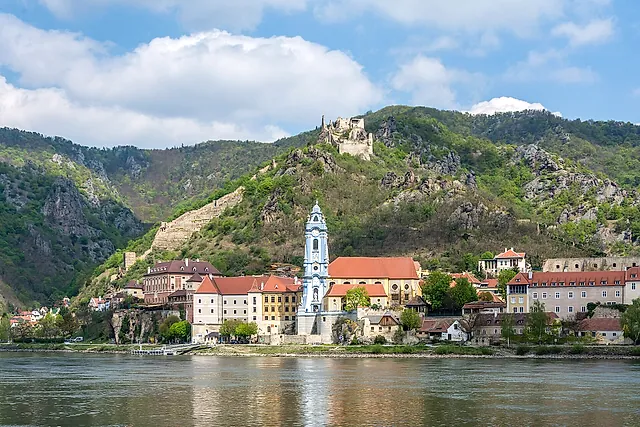 Dürnstein às margens do Danúbio, na Wachau, com a torre azul da igreja da abadia, as ruínas do castelo na colina e o rio em primeiro plano, sob um céu com nuvens brancas