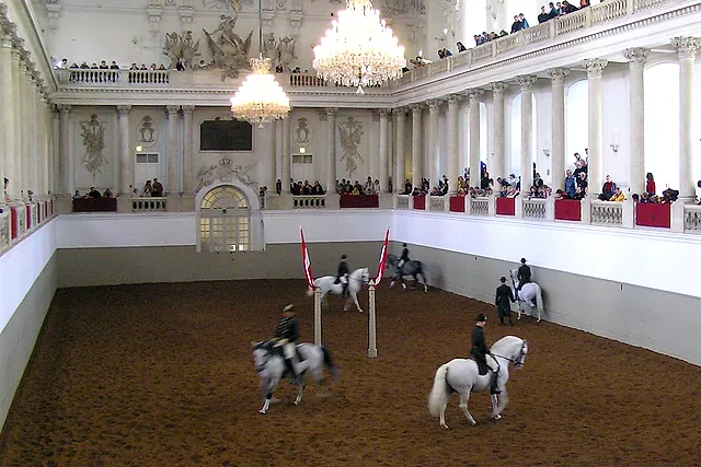 Cavaleiros em cavalos lipizzanos brancos e cinza em uma apresentação no Picadeiro de Inverno da Escola Espanhola de Equitação de Viena, com lustres e o público na galeria