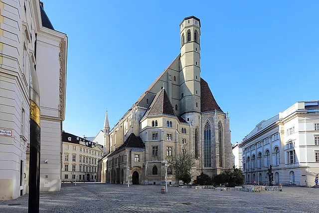A Minoritenkirche gótica, com sua alta torre de pedra, na tranquila Minoritenplatz, sob um céu azul sem nuvens