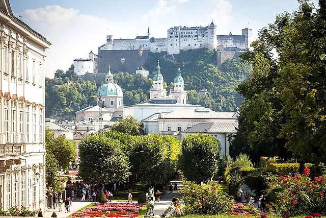 Os jardins de Mirabell em Salzburgo em um dia de sol, com os canteiros de flores, uma ala do palácio à esquerda, as cúpulas da catedral e a fortaleza de Hohensalzburg na colina ao fundo
