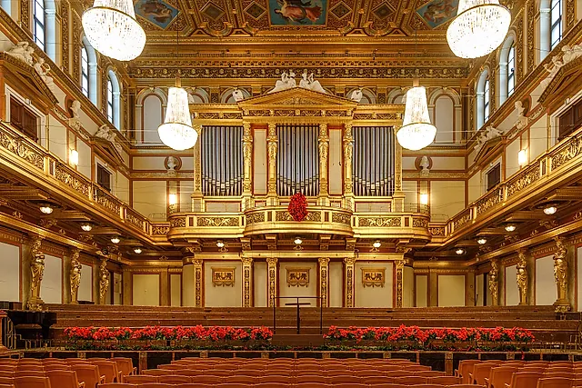 Uma sala de concertos dourada de Viena, vazia, com o grande órgão, lustres de cristal, teto pintado e flores vermelhas ao longo da borda do palco