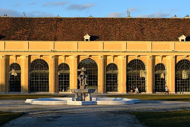 A longa fachada amarela da Orangerie nos jardins do Palácio de Schönbrunn, com as altas janelas em arco e uma fonte à frente, sob um céu azul
