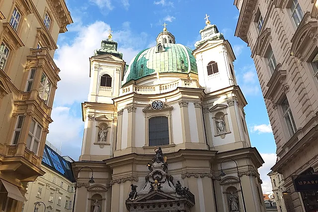 A fachada barroca da Peterskirche, com a cúpula verde e duas torres, vista de uma rua estreita do centro de Viena, sob um céu azul