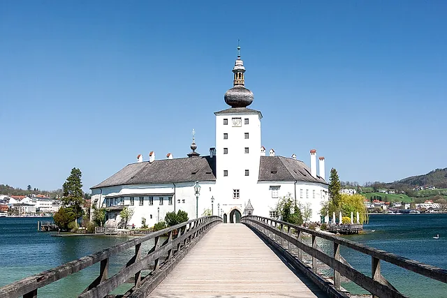 O castelo branco de Ort, com sua torre de cúpula em forma de cebola, em uma pequena ilha do lago Traunsee, alcançada por uma ponte de madeira, sob um céu azul
