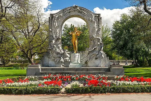 A estátua dourada de Johann Strauss filho tocando violino, emoldurada por um arco de mármore branco, no Stadtpark, com flores vermelhas e brancas na frente e árvores verdes ao redor