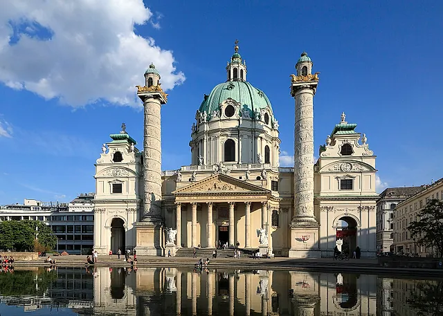 A Karlskirche com sua cúpula verde e as duas colunas com relevos, refletida no tanque da Karlsplatz sob um céu azul