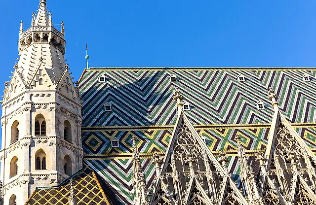 O telhado de telhas vidradas da Stephansdom, com ziguezagues e losangos verdes, brancos, escuros e amarelos, uma das suas torres e os frontões góticos, sob um céu azul