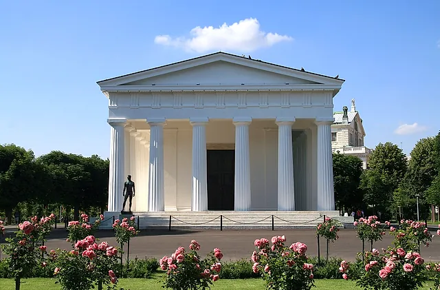 O Templo de Teseu, no Volksgarten, um templo branco com colunas dóricas, com roseiras cor-de-rosa em primeiro plano sob um céu azul